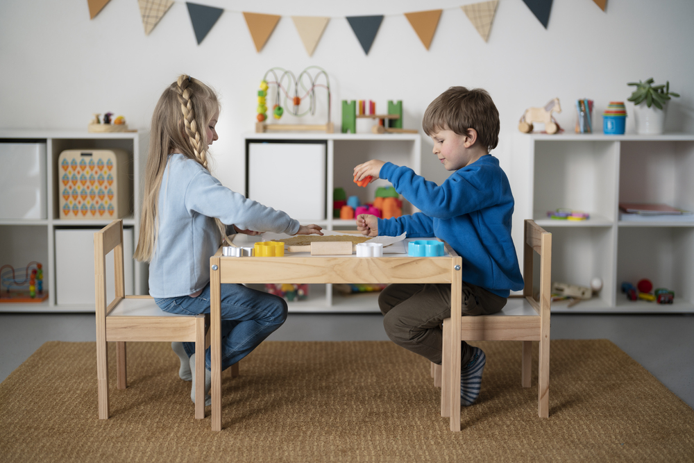 Niños jugando en una mesa infantil de madera en un espacio diseñado para el aprendizaje y la creatividad, resaltando la importancia del mobiliario adecuado en su desarrollo.